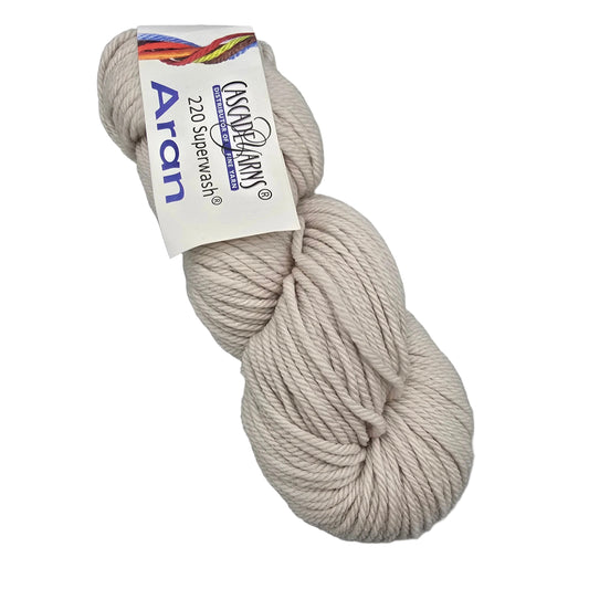 A skein of 220_Superwash_Aran_Yarn_by_Cascade_Yarns in the 272_White_Sand, a light beige color, with label attached in a white background.