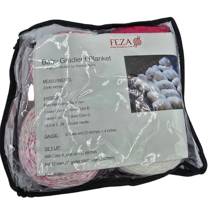 A packaged baby gradient blanket pattern kit by Feza on a white background. The kit is sealed in a clear plastic bag with a black zipper, containing yarn balls in pink, red, and white, along with an instruction card detailing measurements, materials, and setup instructions.