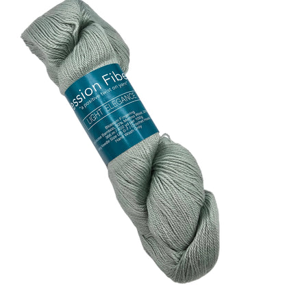 A skein of light sage green Blossom Fingering by Expression Fiber Arts yarn in the color Light Elegance with a teal paper label, photographed on a clean white background.