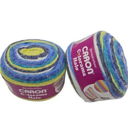 Two skeins of Caron Colorama Halo yarn  in the color Splash a Bluem White and green yarn on a white background. One skein is upright, showing a purple label, and the other is lying on its side, showing the variegated yarn texture.