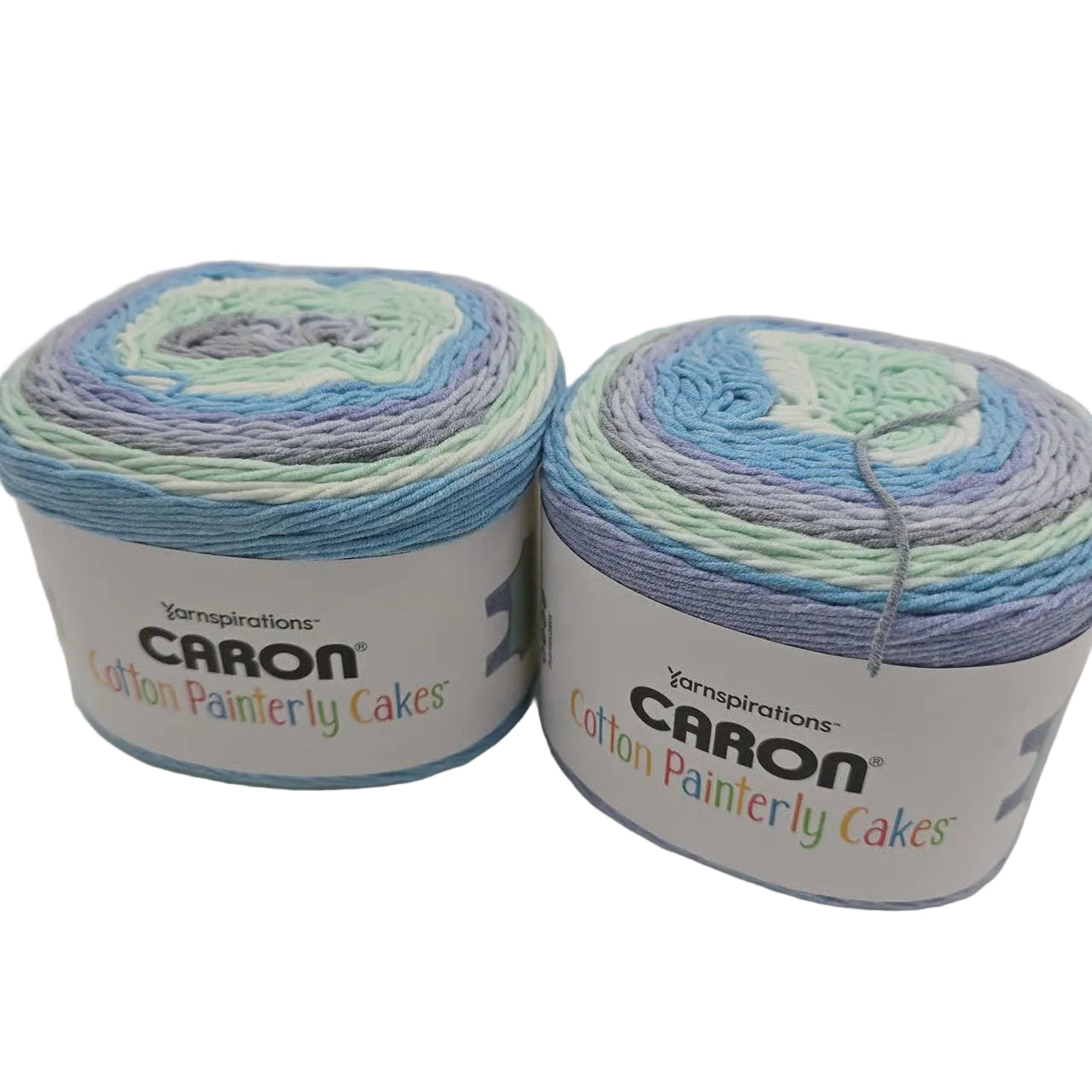 Two Cake of CARON Cotton Painterly Cakes yarn in the color ocean oasis a variegated light blue, mint green, lavender, and white colorway, displayed on a white background.