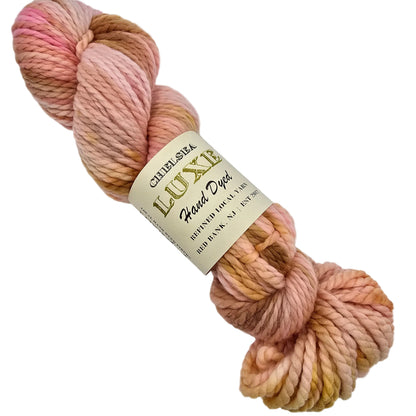 A skein of Chunky_Yarn by_Chelsea_Luxe_in the color Sugar Cookie avariegated yarn with soft pastel shades of pink, peach, beige, and light yellow, wrapped in a cream paper label that reads 'Charleston Luxe Soft Dye' on a white background.
