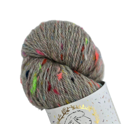 A skein of Corrie_Confetti_by_La_Bien_Aimee_in the color_Greybow_2_Fall_Winter_2021 variegated yarn in a heathered taupe-gray color with colorful flecks, partially wound on a white paper cone with a gold foil sheep outline.