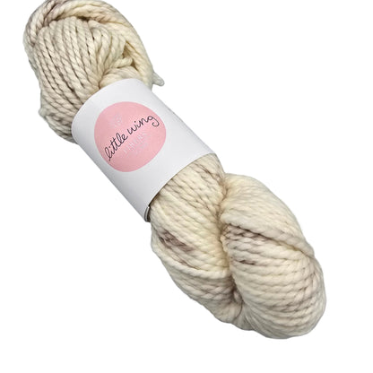A skein of Cloud_Bulky_yarn by_Little_Wing_Fibers in the color Pre-Bake a cream-colored variegated yarn with  a label on a white background.