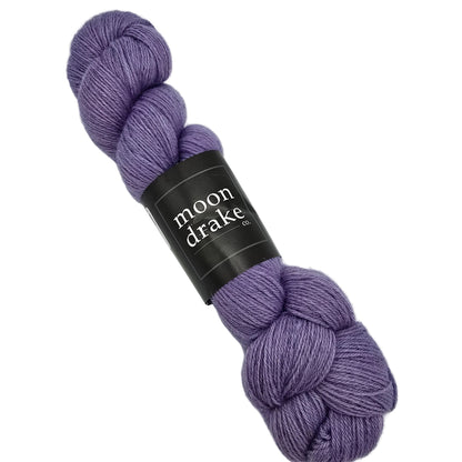 A skein of Corridale Fingering Yarn by Moon Drake Co. in Liliac a dusty lavender-purple color, wrapped with a black paper label, on a solid white background.
