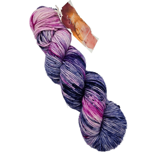 A single skein of variegated yarn with a paper tag, showing a blend of purple, pink, and blue tones, photographed on a light gray background.