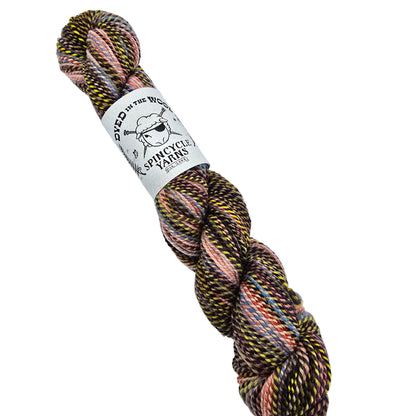 A skein of Dyed_in_Wool_yarn by_Spincycle_Yarns in the color Ghost_Ranch a variegated yarn from featuring a blend of black, pink, yellow, and orange threads, with a white paper label wrapped around the middle.