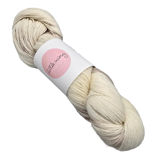 A skein of Eco Sock Yarn by Little Wing Fibers in the color Pre-Bake a cream-colored yarn lightly speckled yarn with label on a white background.