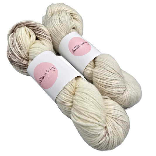Two skeins of Featherlight_DK_yarn by_Little_Wing_Fibersin  variegated cream and beige with labels on a white background.