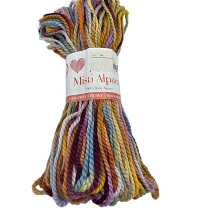 A skein of Misti Alpaca hand-painted chunky yarn with a white paper label, showing a multicolor mix of pastel and earthy tones including purple, blue, yellow, orange, and green on a plain white background.