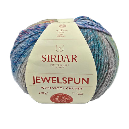 A ball of Jewelspun_with_Wool_Chunky yarn_by_Sirdar_in color 0203_Pearl, wrapped in a cream-colored paper label. The yarn is variegated with shades of blue, purple, pink, and gray on a white background.