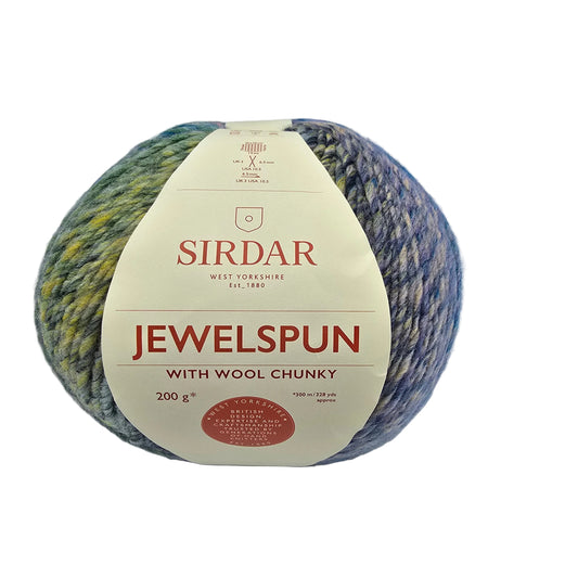 A ball of Jewelspun_with_Wool_Chunky_yarn by_Sirdar in the color Shimmering_Sea_Glass, featuring a cream-colored paper label. The yarn has a variegated color pattern transitioning from olive green and mustard yellow on the left to blue and purple on the right, photographed on a white background.