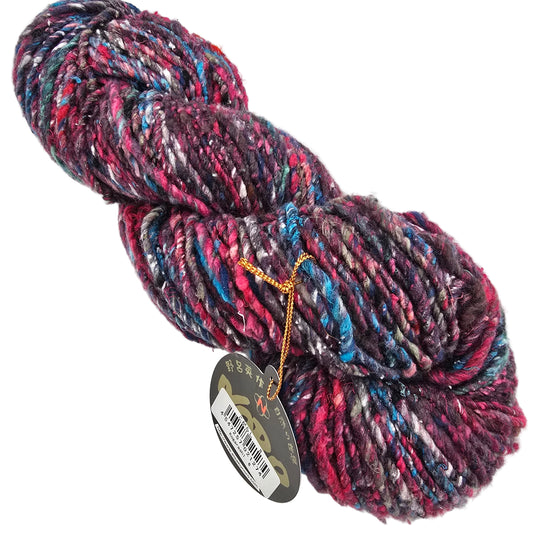 A skein of variegated Kogarashi_yarn by_Noro_-_Color_8  with a marled pattern of pink, purple, blue, and white, featuring a circular tag on a gold string, photographed on a white background.