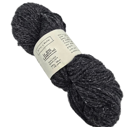A skein of Le_Gros_Lambswool_Yarn_by_Biches_Buches in the color Dark_Grey a dark charcoal gray heathered yarn with a cream paper label wrapped around it, photographed on a solid white background.