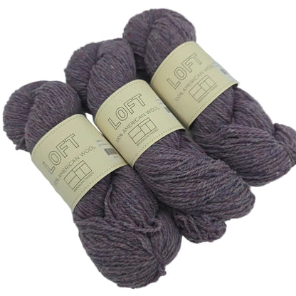 Three skeins of purple variegated Loft by Brooklyn Tweed yarn in the color Blanket Fort with cream paper labels wrapped around them on a white background.