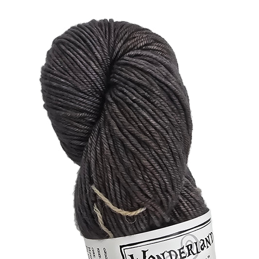 Close-up image of a single skein of Mad Hatter by Wonderland Merino Sport yarn in Tulgey Wood which is a Dark greyish brown color with label on a white background.