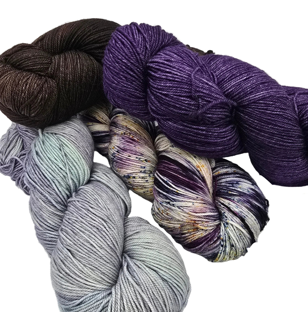 Six skeins of yarn arranged on a white background, including solid colors like deep purple and dark brown, as well as variegated shades of lavender, gray, and purple with small gold and cream flecks. Product tags are partially visible.