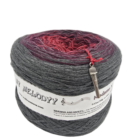 A cake of variegated yarn with a color gradient transitioning from dark gray to deep red and purple, featuring a white paper label and a silver metal tag.