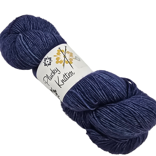 Image of a single skein of Plucky Knitter  Merino, Cashmere, Nylon blend yarn in the color After Dark which is a navy blue with label on a white background.