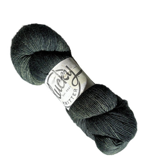 Image of a single skein of Kraken, a dark grey and green, Fingering weight yarn from Plucky Knitter with label on a white background.