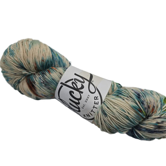 Image of a single skein of yarn from the brand Plucky Knitter in the color Welcome Home which is a cream base with multicolor areas with label on a white background.