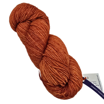 A single skein of Rios_Yarn_by_Malabrio_-in color_016_Glazed_Carrot arust-orange yarn with a white and purple tag, photographed on a white background.