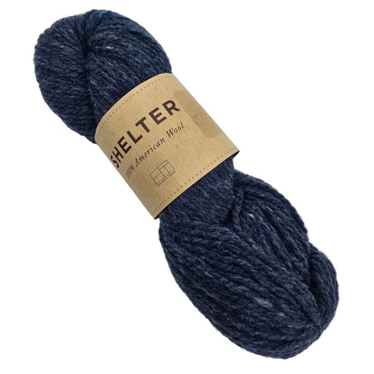 A skein of Shelter by Brooklyn Tweed yarn in Almanac a dark navy blue yarn with a tan paper label that reads 'SHELTER 100% American Wool', photographed on a plain light background.