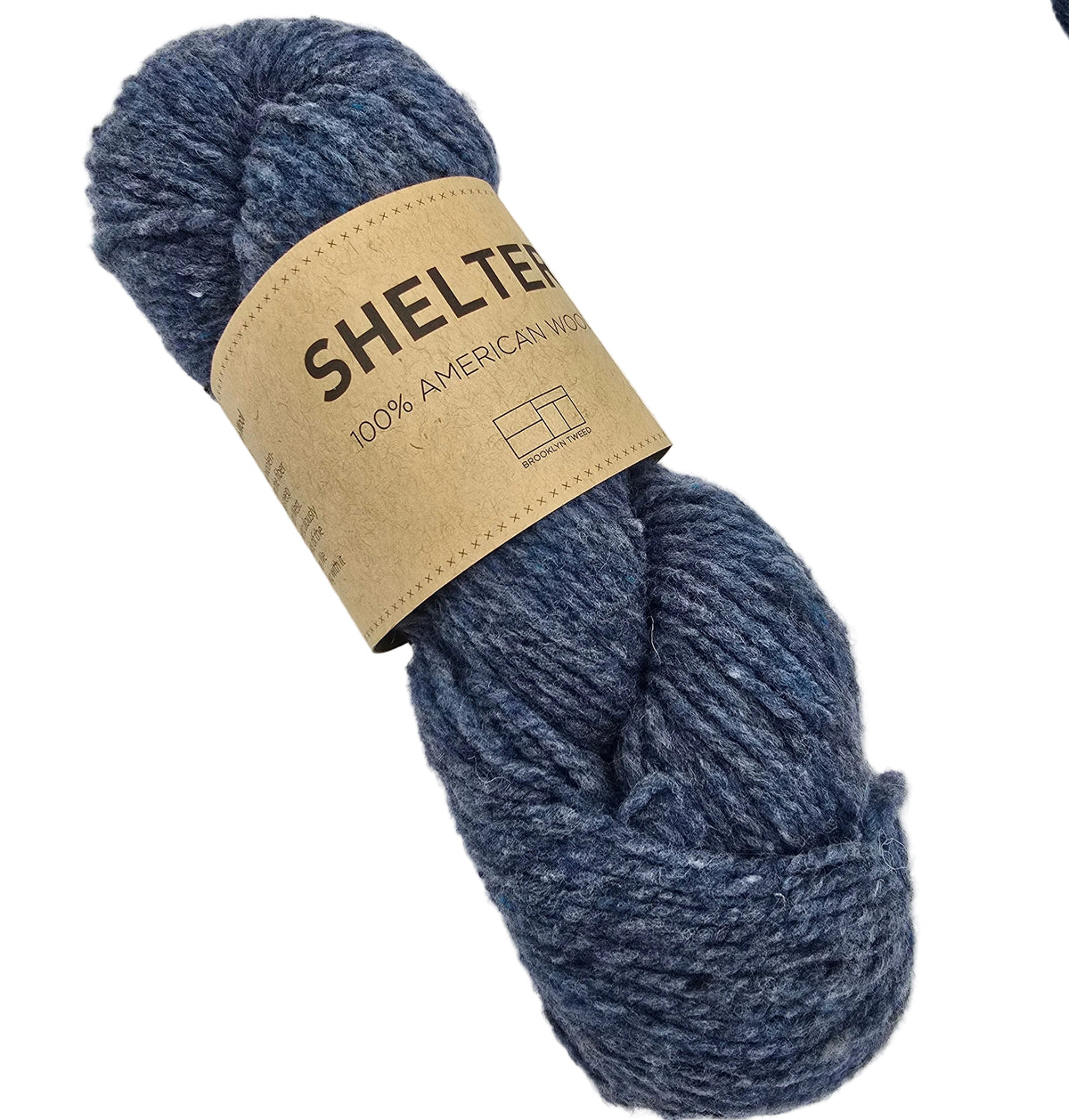 A skein of Shelter by Brooklyn Tweed yarn in the color flannel a heathered blue-gray yarn wrapped with a tan paper label that reads 'SHELTER 100% AMERICAN SILENCE,' photographed on a plain white background.