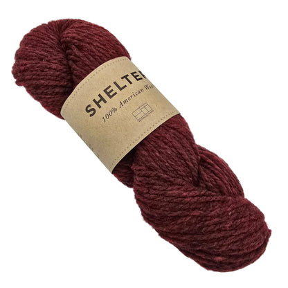 A skein of Shelter by Brooklyn Tweed yarn in the color Long Jonhs a textured yarn in a deep burgundy color, wrapped with a beige paper label that reads 'SHELTER 100% American Wool'.