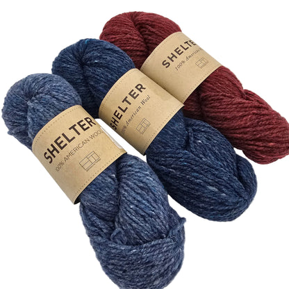 Three skeins of yarn arranged on a light gray background: one burgundy, one heathered navy blue, and one heathered medium blue. Each skein has a tan paper label reading SHELTER.