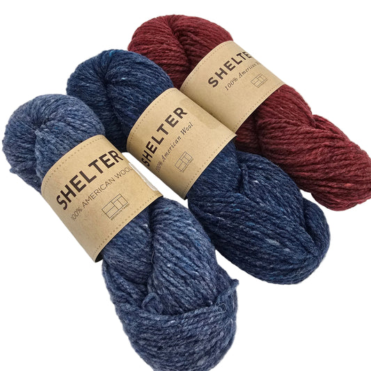 Three skeins of yarn arranged on a light gray background: one burgundy, one heathered navy blue, and one heathered medium blue. Each skein has a tan paper label reading SHELTER.