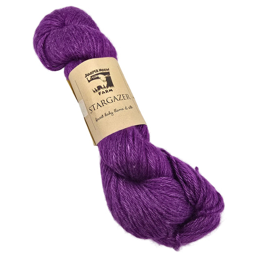 A skein of Stargazer_Yarn by_Juniper_Moon_Farms_- in the color 23 Stargazer a vibrant purple yarn with a kraft paper label photographed on a white background.