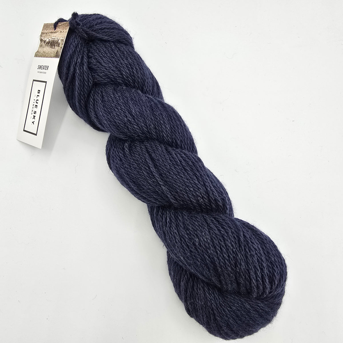 A skein of Sweater Worsted Yarn by Blue Sky Fibers in the color Rainstorm a Navy blue yarn with a paper label attached, photographed on a clean white background.