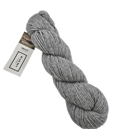A skein of Sweater Worsted Yarn by Blue Sky Fibers in the color Beluga a grey yarn with a paper label attached, photographed on a clean white background.