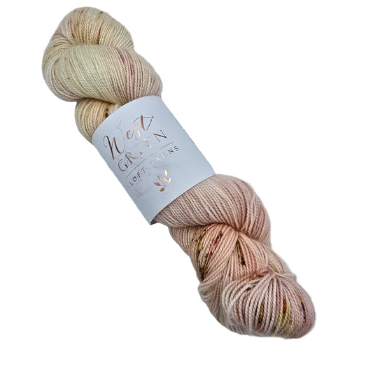 A skein of Swirl_yarn by_West_Green_Loft_Yarns_-in the color Serendipity a variegated cream, peach, and pink colorway with rose gold foil lettering on a white paper label on a whtie background.