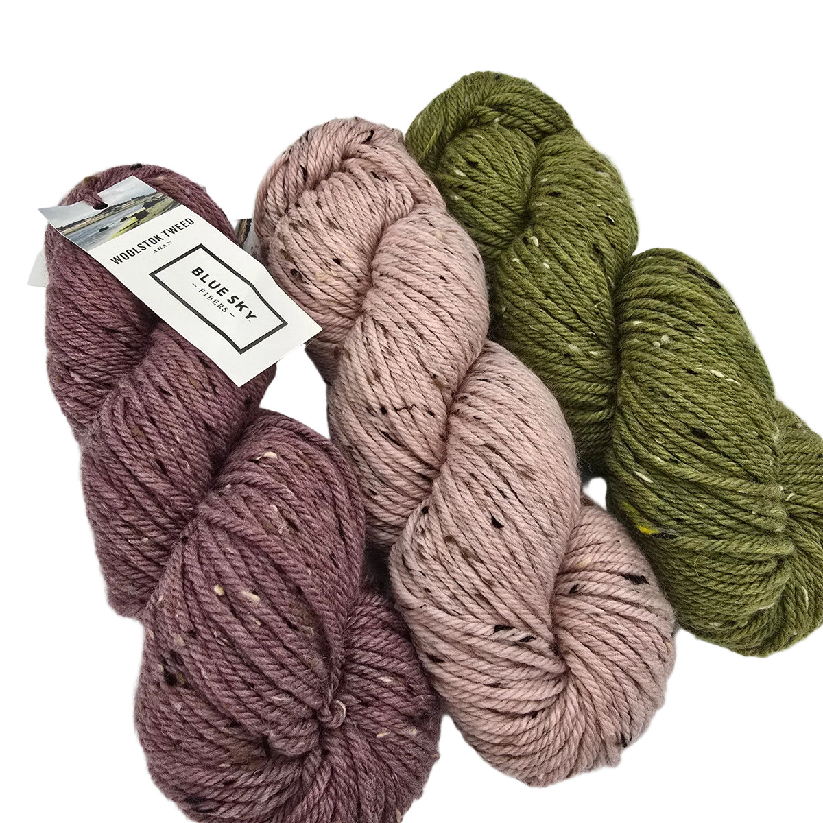Three skeins of Woolstok Tweed Yarn by Blue Sky Fibers arranged on a white background. The yarn comes in three colors: a dusty mauve-purple, a muted dusty pink, and an olive green.