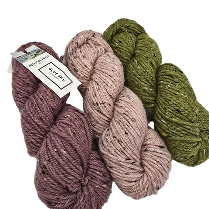 Three skeins of Woolstok Tweed Yarn by Blue Sky Fibers arranged on a white background. The yarn comes in three colors: a dusty mauve-purple, a muted dusty pink, and an olive green.