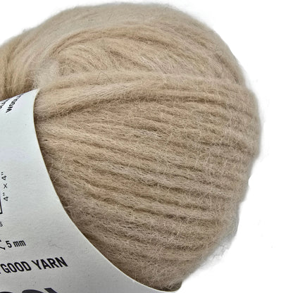 Close-up Image of a single ball of Feeling Good Yarn by Wool And The Gang in the color Blonde Beige with a partially visible label on a white background.
